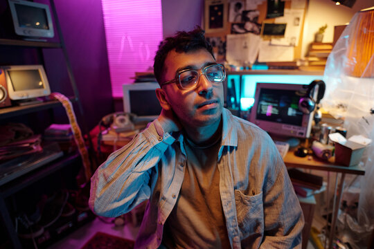 Young male programmer wearing glasses sitting in dimly lit room with retro devices and notice board in background