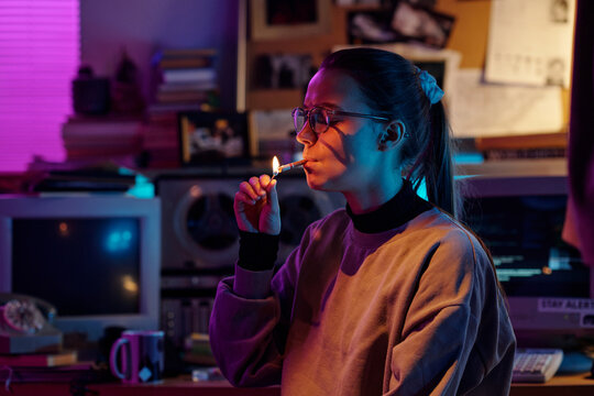 Young woman with glasses and hair in ponytail lighting up cigarette in dimly lit room, medium closeup shot