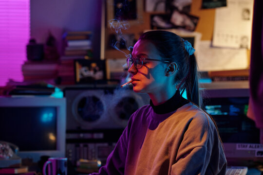 Young Caucasian woman with glasses and hair in ponytail smoking cigarette in dimly lit room using match