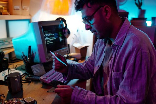 Young male hacker examining three floppy disks while sitting at table with telephone, vintage computers, reel-to-reel tape recorder, ashtray and matchbox on it