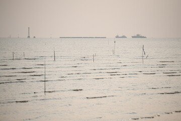 A minimalist seascape under a soft, hazy sky, featuring a vast expanse of calm water with subtle light reflections. Distant maritime structures, including a prominent lighthouse or beacon and a cargo 