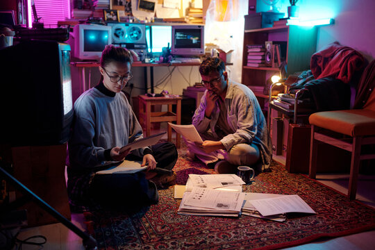 Two diverse young male and female conspiracy theorists studying criminal profiles while sitting on carpet in dark messy room