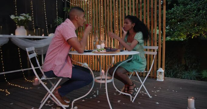 Diverse couple enjoying romantic dinner in garden, surrounded by fairy lights and candles