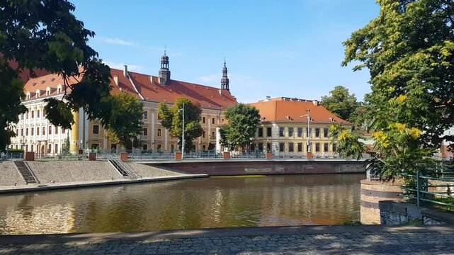 A scenic view of the tranquil Oder River, lined with vibrant green foliage and the quaint, colorful facades of Wrocław's Old Town (Breslau), Poland, basking in the sunlight