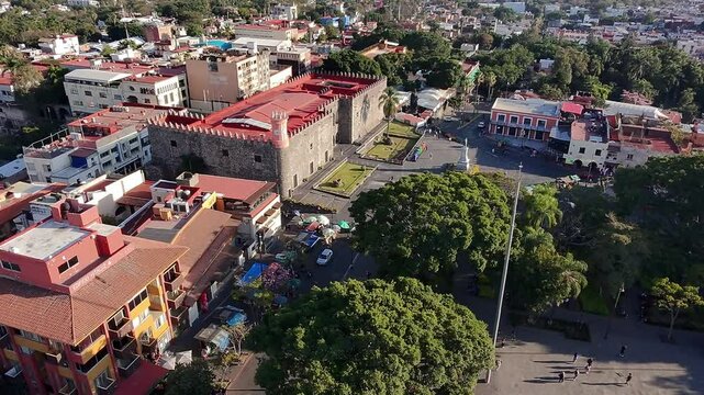 Aerial orbit shot of Cuernavaca city center showing Palacio de Cort&eacute;s, tree-filled Plaza de Armas, colorful Morelos letters, and the Carlos Pacheco monument under clear afternoon light