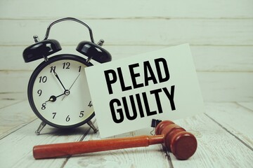 Plead Guilty text on paper card with alarm clock and wooden judge gavel on wooden background