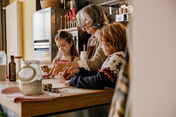 An elderly Caucasian grandmother guides her school-aged grandchildren while baking in a kitchen. They prepare a pastry with utensils and ingredients scattered on the counter.