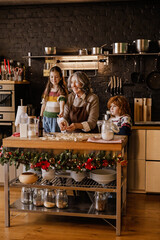 A happy White grandmother in her 60s in an apron cooking cookies with a school-aged granddaughter and a school-aged grandson at a table with ingredients and utensils in the kitchen with black walls