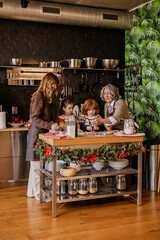A school-aged White brother and sister, making some dough in the kitchen that is decorated for Christmas, together with their mid-30s mother and mid-70s grandmother, at home