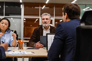 A middle-aged White businessman in his late 50s, sitting among a diverse group of his colleagues at a table in an office room, working on his laptop and listening to them discuss