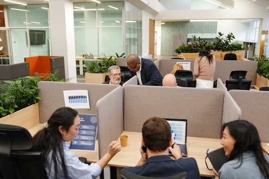 A Black businessman in his late 30s, standing next to a row of cubicles in a spacious office and talking to his middle-aged White male colleague, who sits in one of them