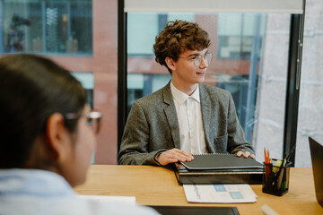 A young White male intern in his late 20s, dressed in a gray business suit, sitting at a table in...