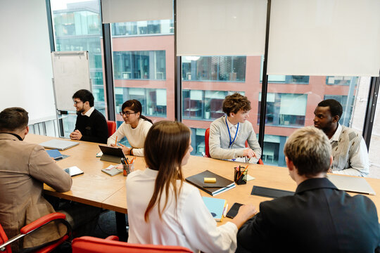 A young White man in his late 20s, talking to his young Black male colleague who sits beside him, as they're sitting at a table in an office meeting room, with their colleagues around them