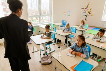 In a sunlit classroom, a Black female teacher stands facing a multiethnic group of elementary...