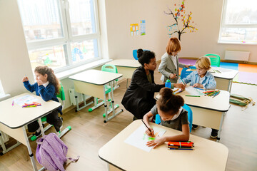 Several diverse elementary school students draw at their desks in a classroom. The room has colorful furniture, decorations, and art supplies. An adult Latino female teacher guides the students.