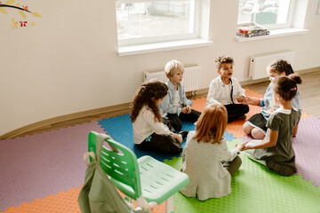 A group of diverse elementary school students participates in a talk while sitting in a circle on colorful floor mats in a classroom with white walls and two windows. A green chair stands beside them.