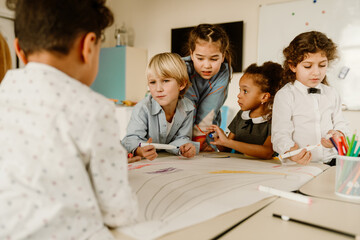Obraz premium A group of multiethnic elementary school children, including a White boy, an Asian girl, and a Black girl, stand at a desk in a classroom, holding markers and drawing on a large piece of paper.