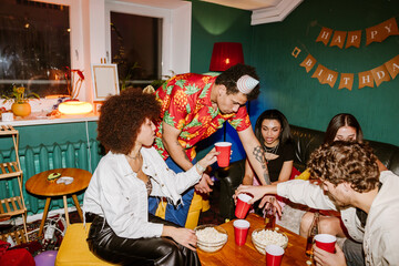 A young Arabic man in his late 20s, standing among a group of his sitting friends in the living room of a cozy home, reaching for something as they're all partying and having drinks together