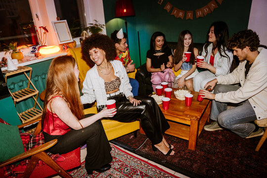 Multiethnic friends enjoy a party while sitting around a wooden table and drinking from red cups as two women in colorful outfits laugh and talk. The women sit on the left in a room with green walls.