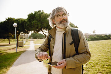 A middle-aged White man in his 50s, with glasses and a short gray beard, standing on a walkway in a grassy park, with a phone in his hand, and looking around, on a sunny day