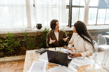 Two diverse women sit at a table with a laptop, documents, and photographs laid out. One wears a brown coat while the other wears a polka dot blouse as they work together and discuss a wedding.