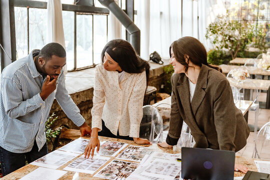 A young Black woman in her late 20s, talking to her Black male fianc?e about some wedding choices, as they stand with their mid-30s White female wedding planner near a table in a spacious hall