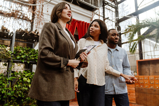 A Black couple in their 20s, a man and a woman, are standing next to each other, holding hands, and talking to a White female consultant, in a botanical garden with large windows, and many plants.