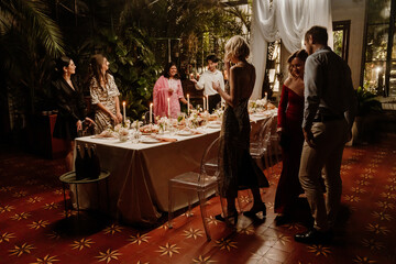A diverse group of young wedding guests, who are all dressed in beautiful clothes, gathering near a long table with petals, plates, and candles on it, getting ready to have dinner with the newlyweds