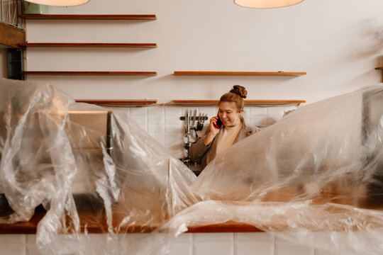 A happy European female adult cafe owner standing at a countertop wrapped in plastic in a cafe with white walls and wooden shelves and talking on the phone, her brown hair tied in a bun