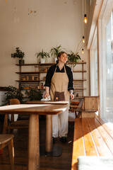 A White woman in her 20s with reddish hair tied back, wearing a black shirt, beige apron, and white pants, stands wiping a wooden table with a white cloth in a sunlit cafe near a large window.