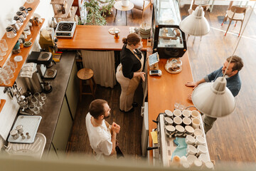 An overhead view of a young White woman in her 20s, working behind the counter in a coffee shop, accepting an order from a mid-30s White man, as her older White male colleague is beside her