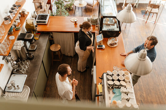 An overhead view of a young White woman in her 20s, working behind the counter in a coffee shop, accepting an order from a mid-30s White man, as her older White male colleague is beside her