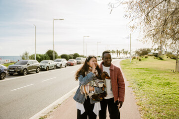 A young Asian woman in her 20s, calling out to someone while holding her dachshund dog in her hands, as she walks with her mid-20s Black male friend away from a road towards a park, on a sunny day