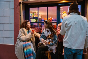 A young Asian woman in her 20s, holding her dachshund dog in her lap and chatting with her young Latina female friend, as they sit outside of a bar in the evening,drinking, with two men beside them