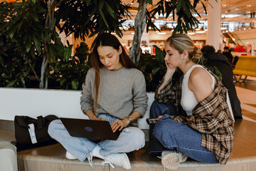 A young, blond White woman in her late 20s, sitting at an airport bench with her legs crossed and a...