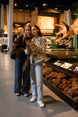 Two young White women stand near a large dinosaur skeleton in a museum. Exposed beams and rustic walls are in the room. The women look at the phone in the hands of the woman in the blue jeans.