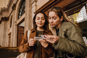 Two young White women in their late 20s, a blond, and a brunette, looking at their train tickets with a happy look on their faces, as they stand inside of a spacious hall of the train station