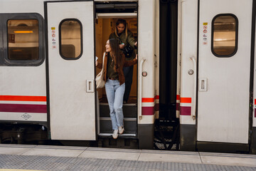Two young Caucasian women exit a train carriage, one with long brown hair wears a brown jacket and light blue jeans, while the other walks behind her in a green jacket and blonde hair.