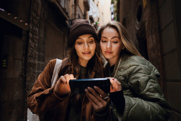 In a narrow alley, two young White women stand, one wearing a brown jacket and beanie and the other...