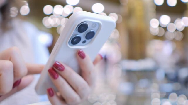 Woman is holding a white iPhone with a pink manicured hand. She is looking at the screen and she is texting