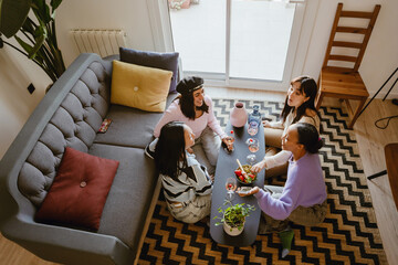 Four diverse women wearing casual clothes gather around a coffee table in a living room, enjoying...