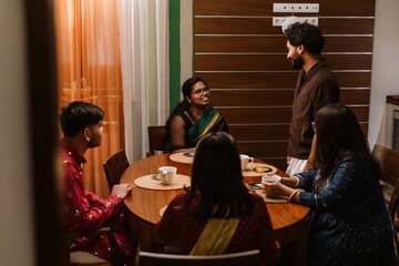 A young Indian man in his late 20s, standing next to the table where his siblings are having tea, talking to his mid-30s sister who sits at the head, in a cozy home