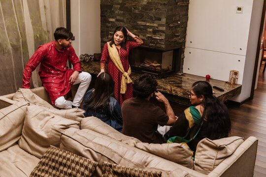 An adult Indian man and a woman wearing traditional Indian attire stand near a couch in a living room with a stone fireplace and white walls. Other Indian friends sit on the couch and talk.