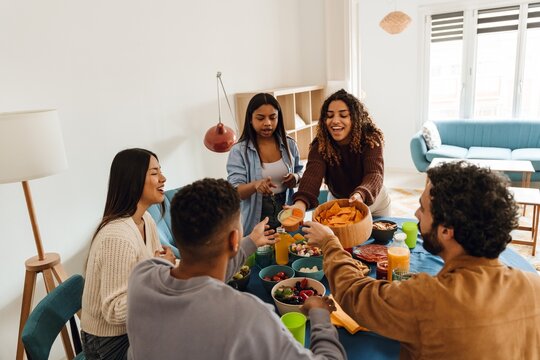 Five diverse friends, three women, and two men, sit and stand around a table enjoying snacks and drinks together. A blue sofa and modern furnishings improve the setting for their casual meal.