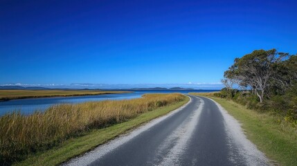 Fototapeta premium Scenic coastal road curving beside calm river under a vibrant blue sky.