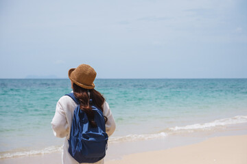 Rear view of a female traveler with hat and backpack standing on the beach by the sea