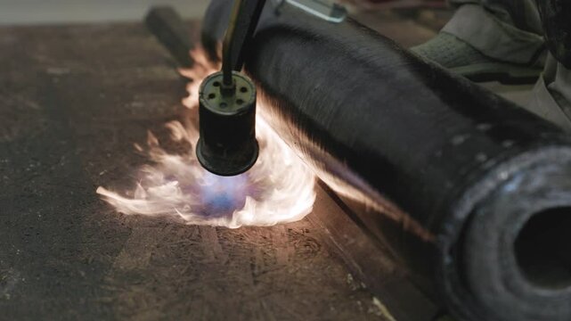 Close-up of a worker using a gas torch to apply heat to a rolled bitumen membrane, sealing it to a rooftop surface in a waterproofing construction process