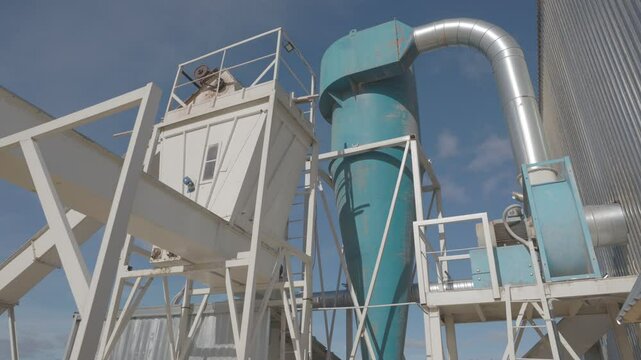 An outdoor view of an industrial cyclone separator with ductwork and conveyor system under a clear blue sky, used for air filtration or material processing
