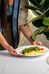 A person serves a freshly made omelette with crispy bacon, arugula, and cherry tomatoes on a plate. The cozy scene includes a warm-toned setting with a soft cardigan and a plant in the background.