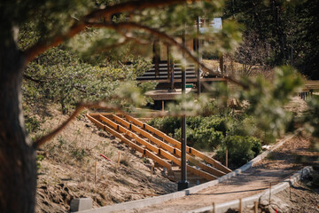 Wooden stairs under construction on a forested hillside lead toward a cabin in the background, partially obscured by tree branches in the foreground..